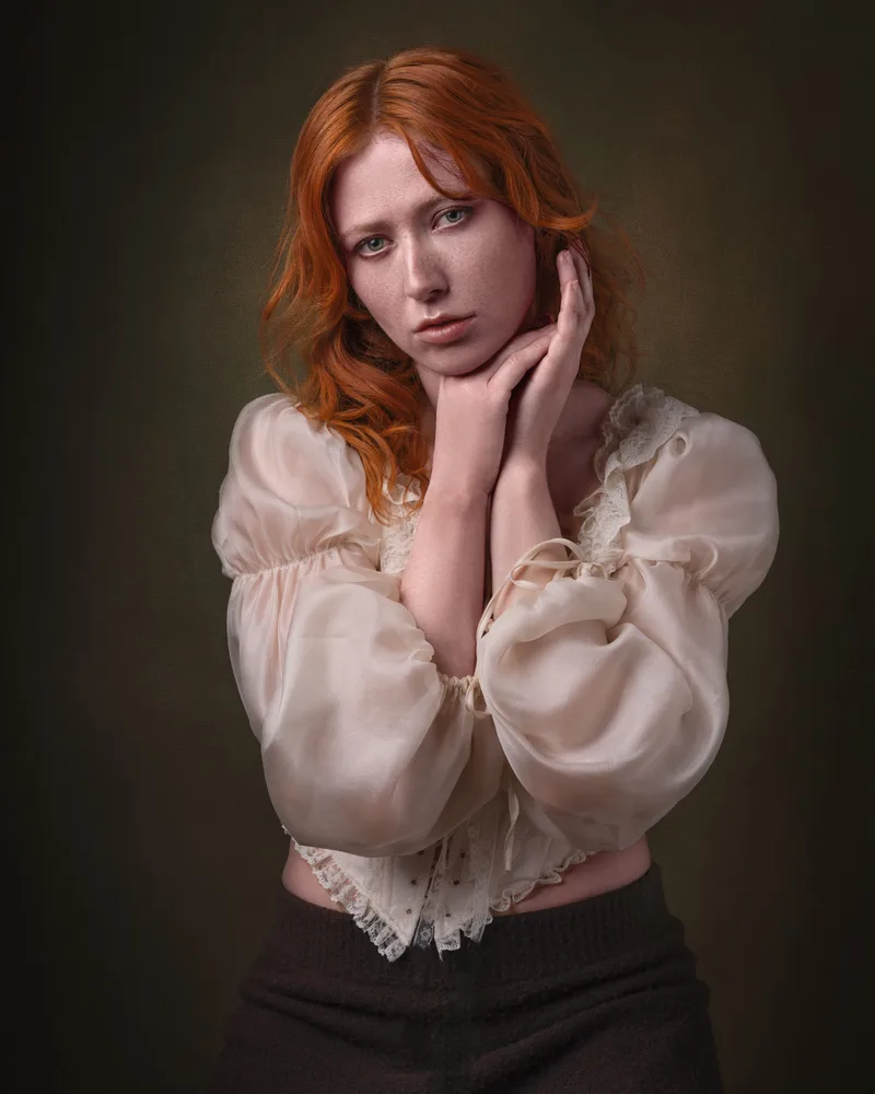 Woman with red hair wearing a cream blouse with lace trim and dark skirt, hand raised to face, posed against a dark olive