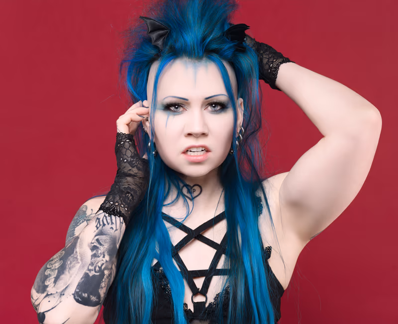 Person with blue and black spiked hair, tattooed arm, and black lace gloves poses with hand raised to head against a deep