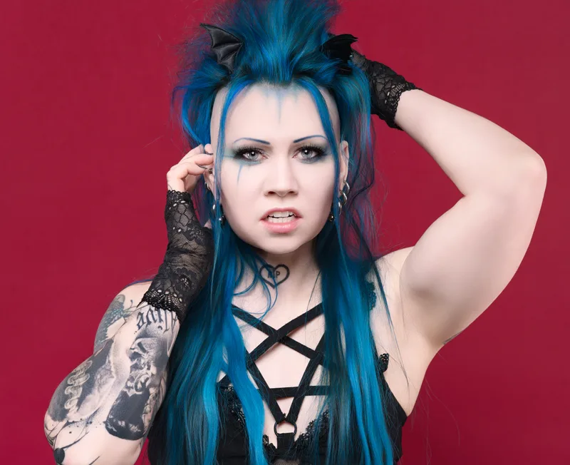 Person with blue and black spiked hair, tattooed arm, and black lace gloves poses with hand raised to head against a deep