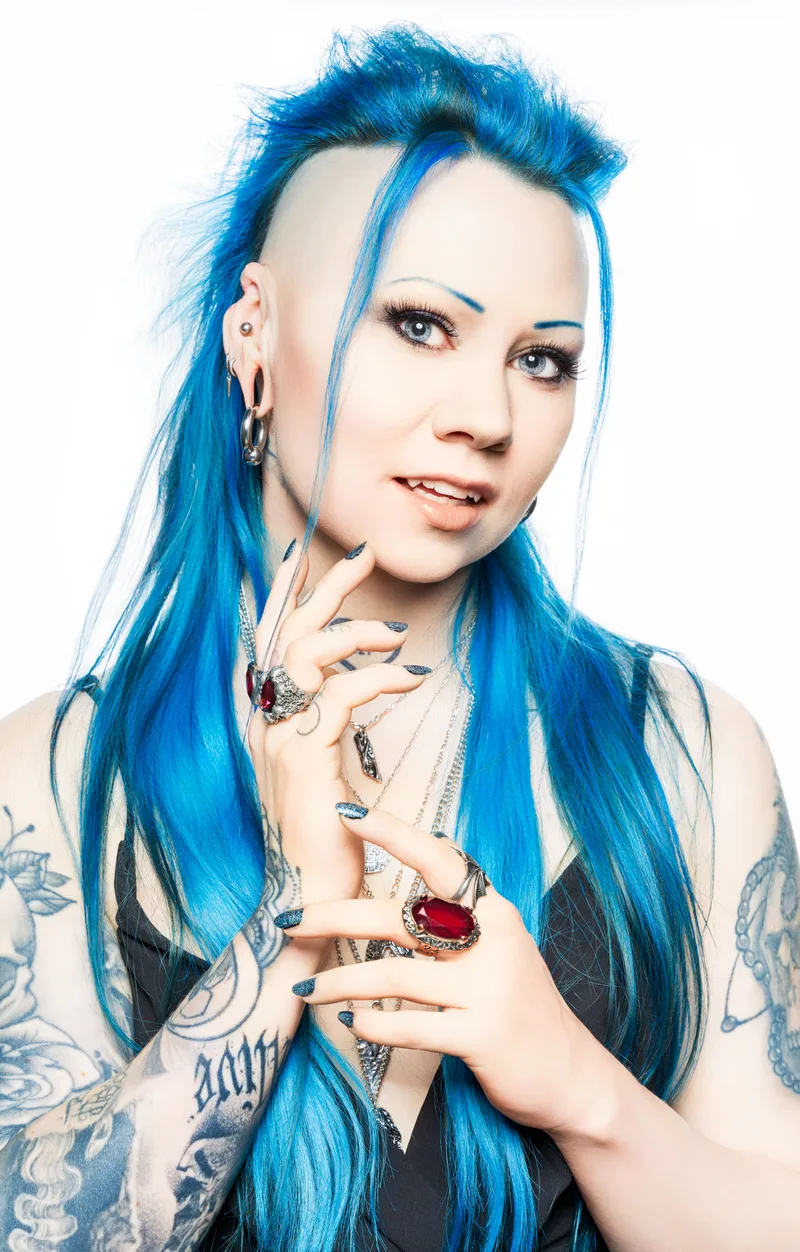 Person with bright blue hair styled with volume on top, blue eyebrows, and multiple piercings wearing silver and red rings,