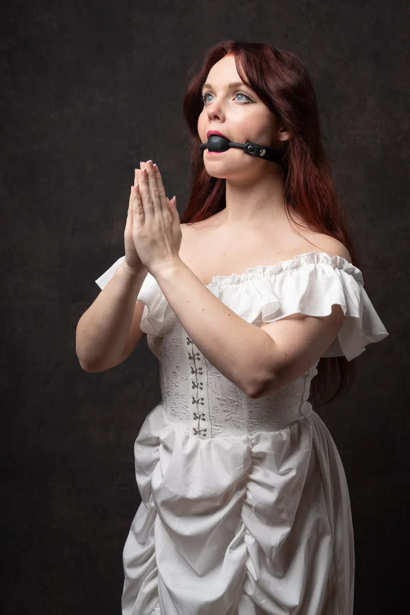 Person with long burgundy hair wearing a white corset dress and black mouth gag, hands held in prayer position, against dark