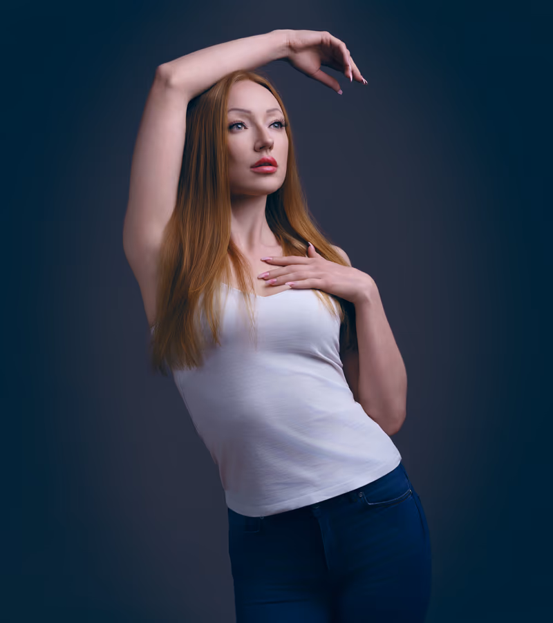 Woman with long reddish-brown hair wearing a white tank top and dark jeans, posing with one arm raised above her head and