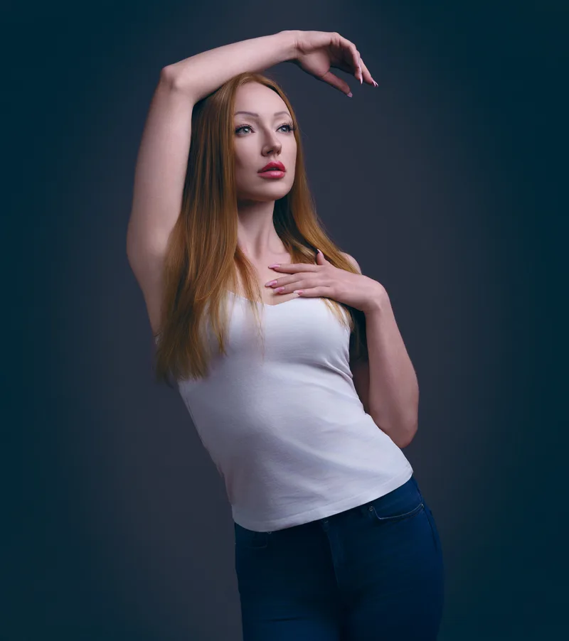 Woman with long reddish-brown hair wearing a white tank top and dark jeans, posing with one arm raised above her head and