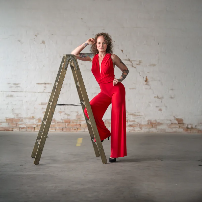 Woman in red halter jumpsuit leaning against wooden ladder with hand to head in industrial space with weathered white and