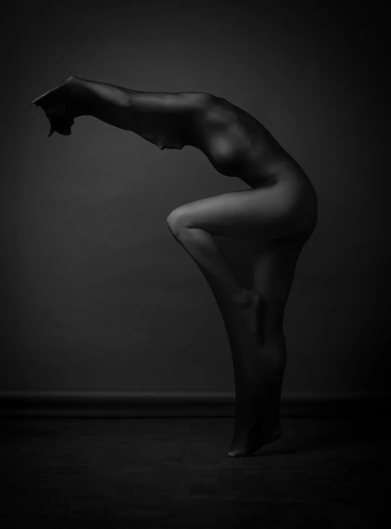 Silhouetted figure in a forward bend pose against a dark gray background with dramatic lighting emphasizing body contours.