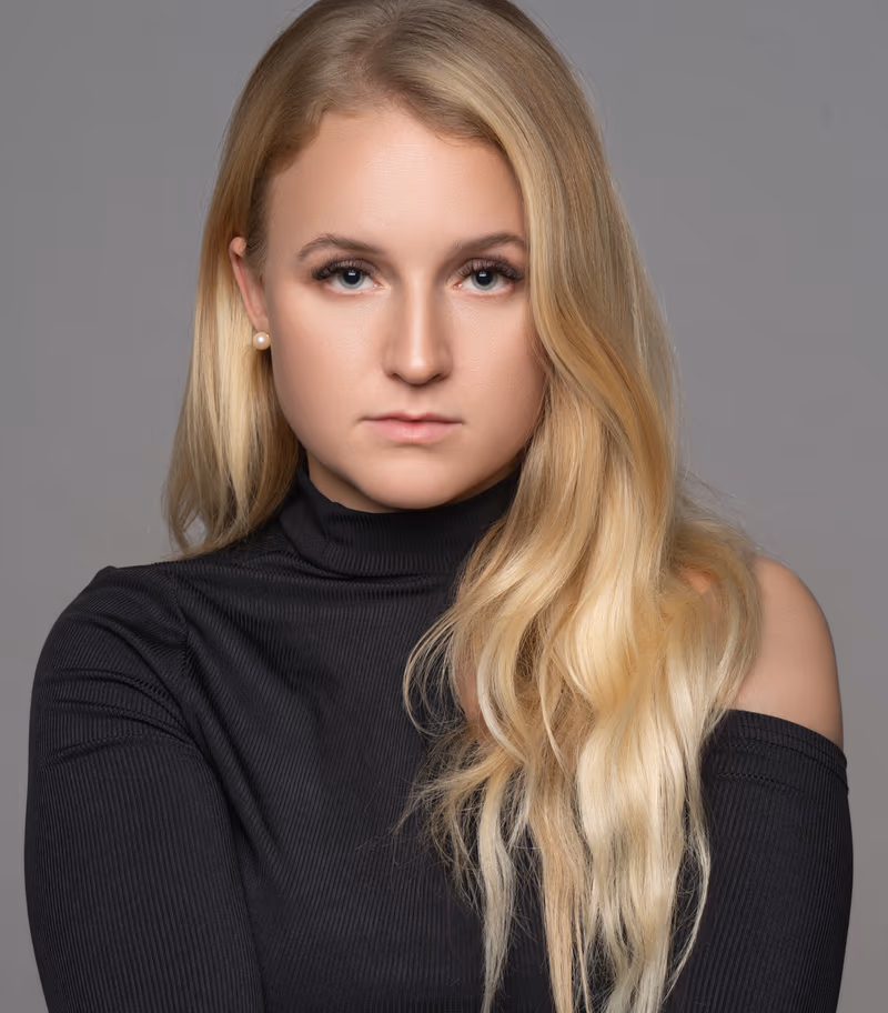 Woman with long blonde hair wearing a black off-shoulder turtleneck, facing camera against neutral gray background with even