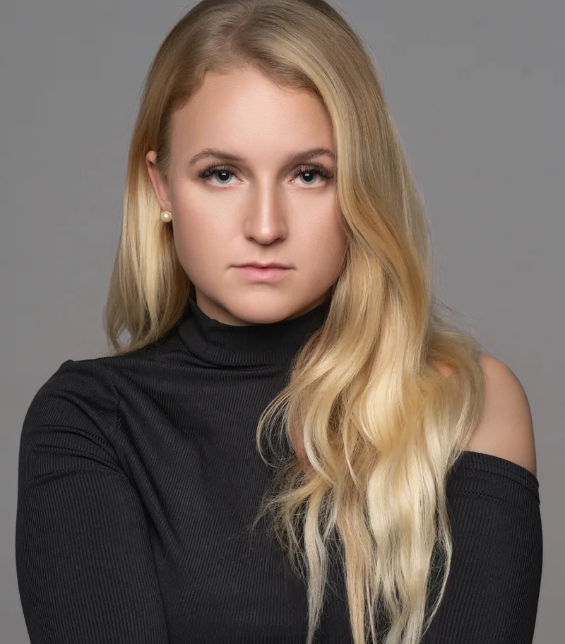 Woman with long blonde hair wearing a black off-shoulder turtleneck, facing camera against neutral gray background with even