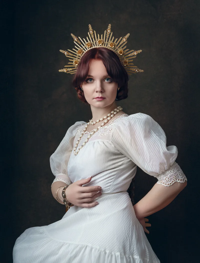 Woman with reddish-brown hair wearing a white pleated dress with puffed sleeves, pearl necklace, and an ornate gold sunburst