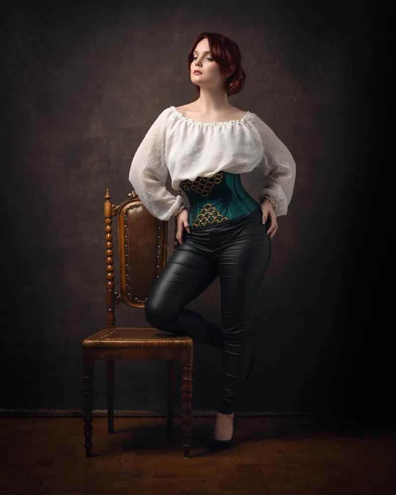 Woman with reddish-brown hair wearing a white peasant blouse, teal corset, and black pants, standing with one foot on a